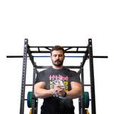 A focused man stands before a weightlifting rack, clapping Bells of Steel Mighty Grip Workout Chalk from his hands. He wears a black tee with colorful text, ready for exercise against a plain white background.