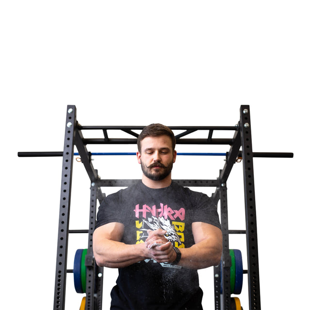 A focused man stands before a weightlifting rack, clapping Bells of Steel Mighty Grip Workout Chalk from his hands. He wears a black tee with colorful text, ready for exercise against a plain white background.