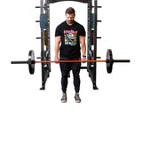 A man stands before a squat rack gripping a loaded barbell, his hands coated in Bells of Steel Mighty Grip Workout Chalk as he prepares to lift, dressed in a black t-shirt, dark pants, and sneakers.