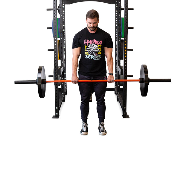 A man stands before a squat rack gripping a loaded barbell, his hands coated in Bells of Steel Mighty Grip Workout Chalk as he prepares to lift, dressed in a black t-shirt, dark pants, and sneakers.