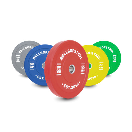Five Bells of Steel Coloured Bumper Plate Sets (15, 25, 35, 45, and 55 lbs; gray, blue, red, yellow, green) are arranged upright in a row. Each plate features a durable stainless steel insert ring.