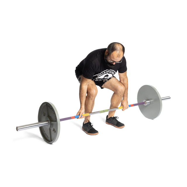 In a gym setting, a man with glasses and sneakers prepares to lift the Bells of Steel Hydra Seven Ring Bar, wearing a black t-shirt and shorts. He showcases his weightlifting prowess as he grips the barbell featuring its multi-colored grips, ready to demonstrate his skill with precision.