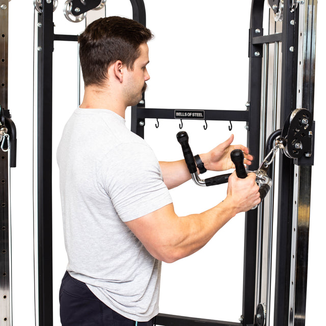 A man wearing a gray T-shirt prepares for his workout using the Bells of Steel Multi Purpose Close Grip Cable Attachment. He firmly grips its rubberized handles, ready to begin his exercise on the cable machine against a plain white background that accentuates his concentration.