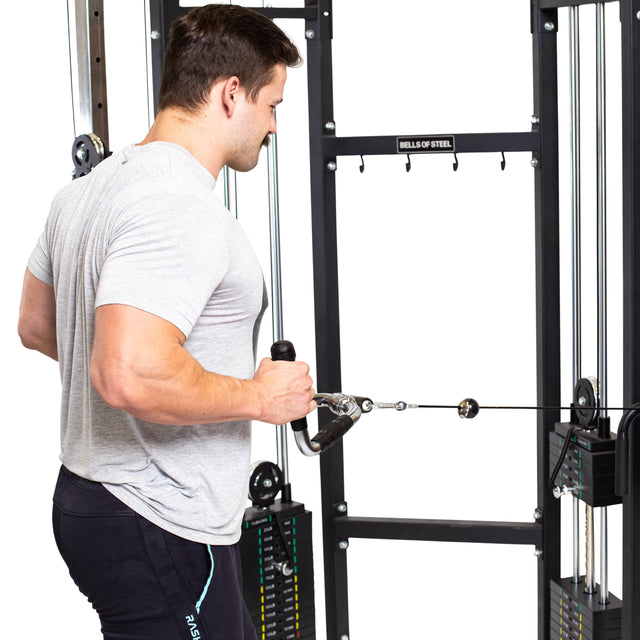 A person in a gray T-shirt is working out at the gym, performing a tricep pushdown exercise with a neutral grip using the Bells of Steel Multi Purpose Close Grip Cable Attachment. This attachment comes with rubberized grips for added comfort. The machine they're using includes metal plates for adjustable weights.