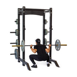 Dressed in black athletic wear and a cap, an individual confidently performs a squat with a barbell using the Hydra Collegiate Power Rack Builder (3" x 3", ⅝" Holes) from Bells of Steel, demonstrating the versatility of its modular components. Multiple weight plates are neatly organized on the sides of the rack against a plain white background.