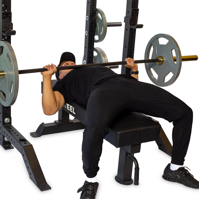 Wearing a black outfit, an individual performs a bench press on a weight bench, utilizing the Bells of Steel Hydra Collegiate Power Rack Builder (3" x 3", ⅝" Holes) for support. They grasp the barbell with both hands, preparing to lift amidst the plain white background, which highlights the focus on this strength exercise.