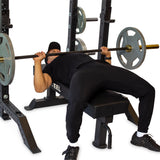 In a gym setting, a person clad in black clothing uses the Hydra Collegiate Power Rack - Prebuilt (3" x 3", ⅝" Holes) by Bells of Steel to perform a bench press, lying on the bench and lifting a barbell with weight plates. The individual appears focused on the exercise.