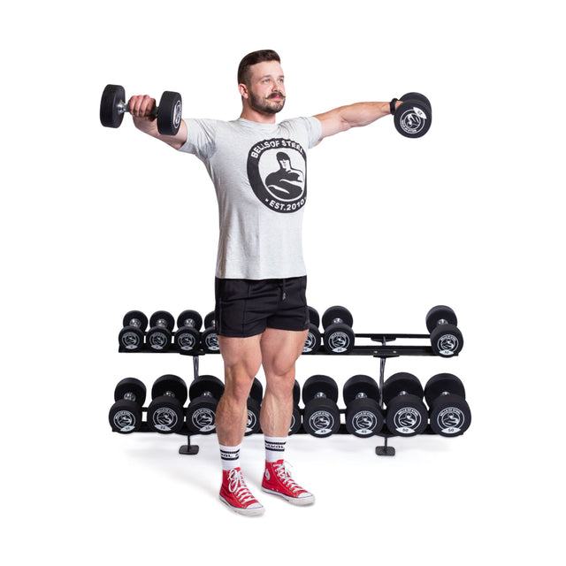 A man is using Bells of Steel Urethane Dumbbells for a lateral raise. He sports a gray T-shirt, black shorts, red high-top sneakers, and striped socks. A rack of various commercial-grade weights stands behind him.