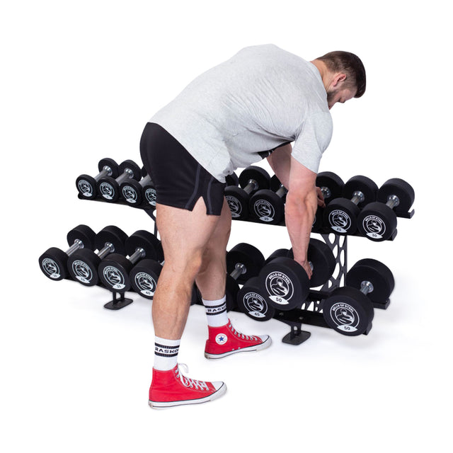 Wearing a gray t-shirt, black shorts, and red sneakers, a person grabs a Bells of Steel Urethane Dumbbell from a rack with multiple weights against a white background.