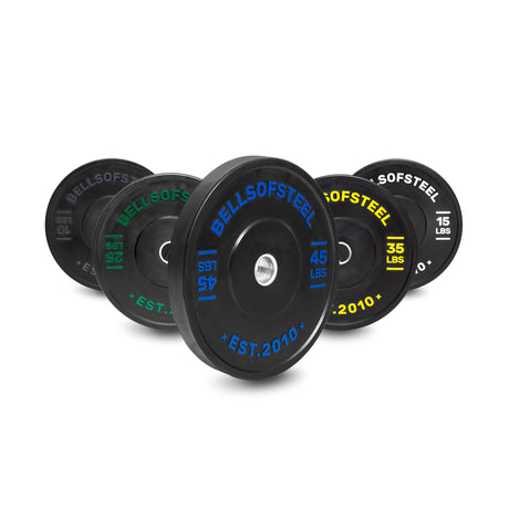 Five Bells of Steel Dead Bounce Conflict Bumper Plates (55lb, 45lb, 35lb, 25lb, 15lb) with colored labels—blue, green, yellow, white—stand upright in a row; each plate features a stainless steel insert and accurate weight markings.