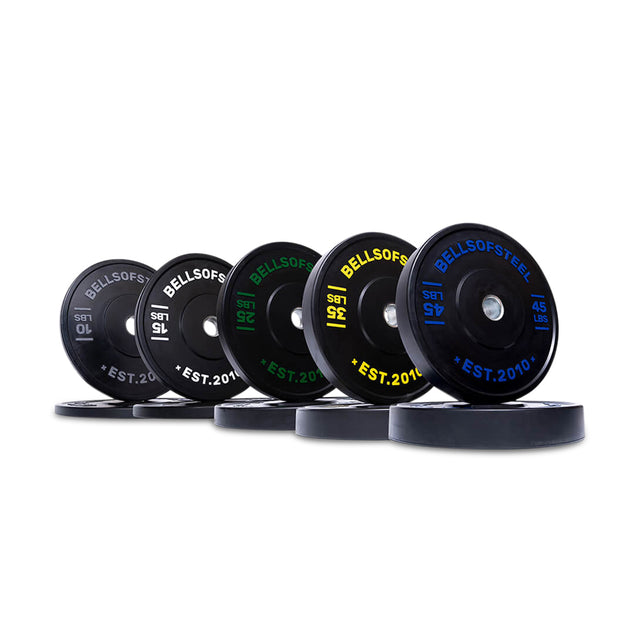 A row of Bells of Steel Dead Bounce Conflict Bumper Plates stands upright, displaying vibrant colored labels for weights in kg and lbs. These plates have a stainless steel insert and are made from durable virgin rubber for optimal performance.