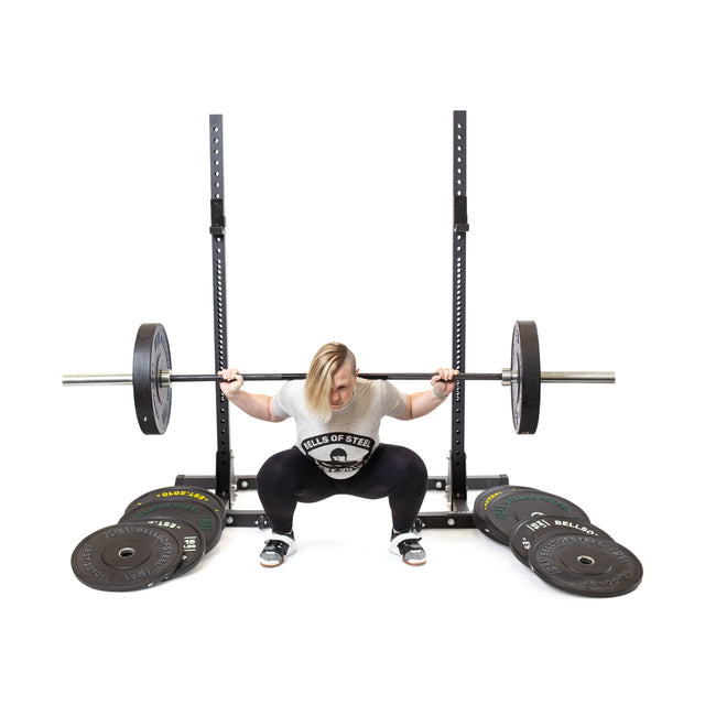 A person in a Bells of Steel shirt squats with a barbell using Dead Bounce Conflict Bumper Plates. They're near a squat rack, with extra weights on the floor.