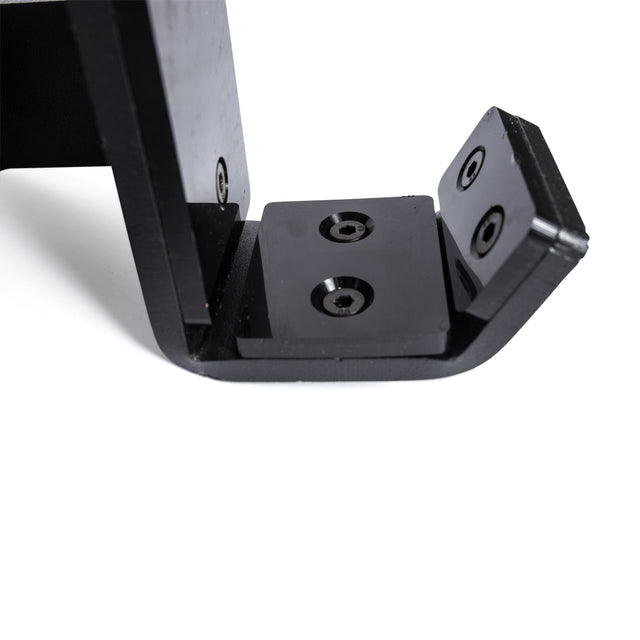 A close-up of the Bells of Steel Standard J-Cups shows its black metal bracket with screw holes and an L-shaped design, making it ideal for a power rack. This sturdy mounting component, positioned on a white surface, is perfect for supporting heavy weights and features added UHMW protection.