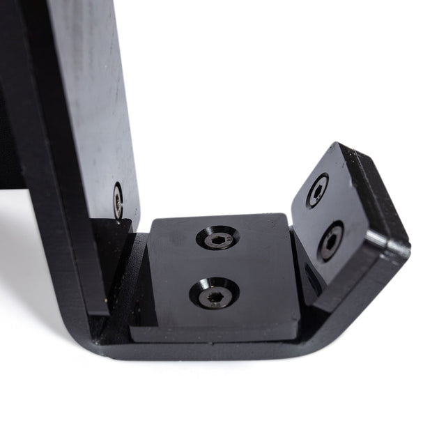 Close-up of the Bells of Steel Standard J-Cups, a black, L-shaped metal bracket with three screw holes, attached to a flat surface. The sleek, glossy finish resembles the robust accessories on a power rack. It's shown from an angle that highlights the polished corners and edges.
