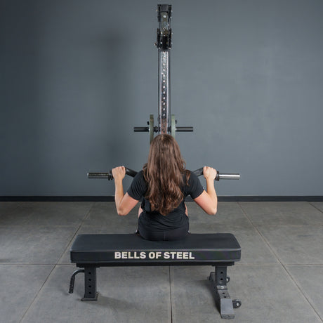 A person with long brown hair, seen from behind, uses the Bells of Steel Fat Bar - Olympic Curl Bar Cable Attachment for a lat pulldown, challenging their grip strength while seated in the gym.