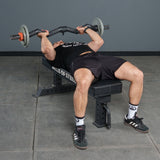 A person is bench pressing with the Bells of Steel Fat Bar - Olympic Curl Bar Cable Attachment in a gym, wearing a black shirt and shorts, white socks, and black athletic shoes to enhance grip strength.