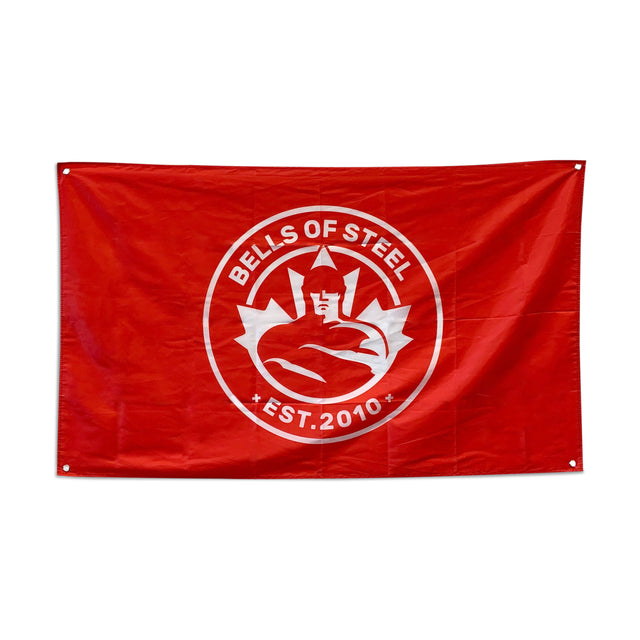 The Bells of Steel flag is a striking addition to any gym decor, featuring a red background with a white circular emblem depicting a stylized muscular figure lifting a barbell. The design includes the text "BELLS OF STEEL" and "EST. 2010." Two eyelets are located on the left corners, making it perfect as a home gym flag.