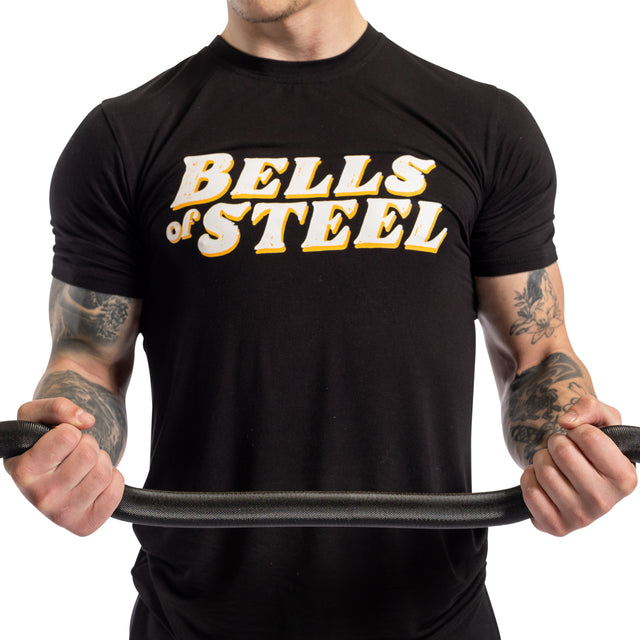 A person wearing a black Bells of Steel T-shirt grips the EZ Curl Bar (54.5”) with both hands, their tattooed arms highlighted against a plain white background, showcasing strength and arm definition.