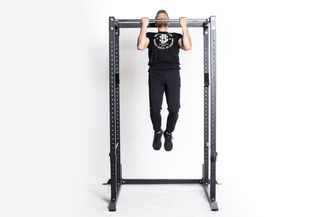 A person wearing a black t-shirt and pants performs a pull-up on a metal pull-up bar stand, which is part of the Residential Power Rack by Bells of Steel, set against a white background.