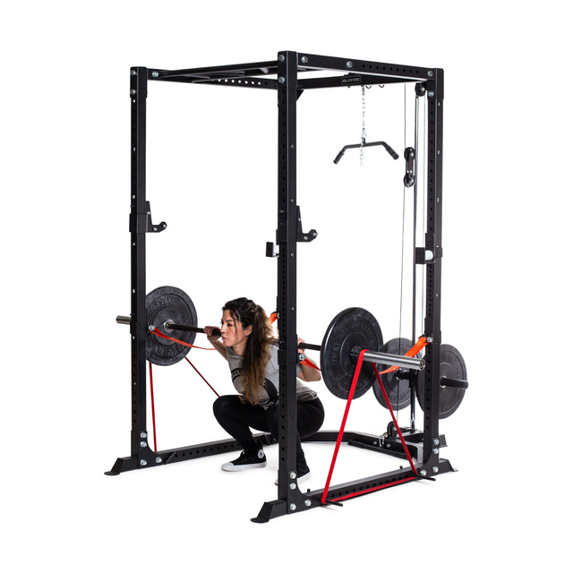 A person squats inside the Bells of Steel Light Commercial Power Rack, securely holding a barbell with weights on each side. The equipment features resistance bands and a pull-up bar, with safety straps ensuring stability. The background is plain white.