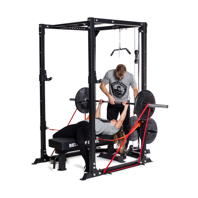A person bench presses a barbell inside the Bells of Steel Light Commercial Power Rack (2.3" x 2.3", ⅝" holes) as another spots them. Both wear gray shirts; resistance bands and safety straps are attached. The background is plain white.