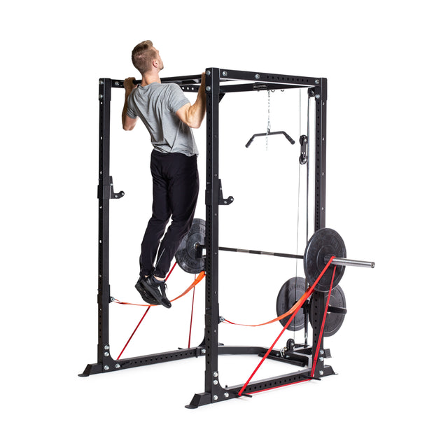 A man performs pull-ups on a Bells of Steel Light Commercial Power Rack, which is equipped with a barbell, weight plates, and vibrant red resistance bands. He is dressed in a gray t-shirt and black pants as he trains in the well-lit indoor gym space.