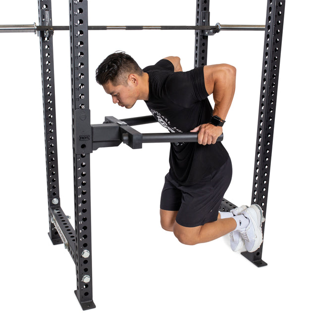 While wearing a black t-shirt, black shorts, and white sneakers, a person performs tricep dips with their knees bent and elbows at a perfect 90-degree angle using the Bells of Steel Y Dip Bar Rack Attachment securely fastened to the power rack.