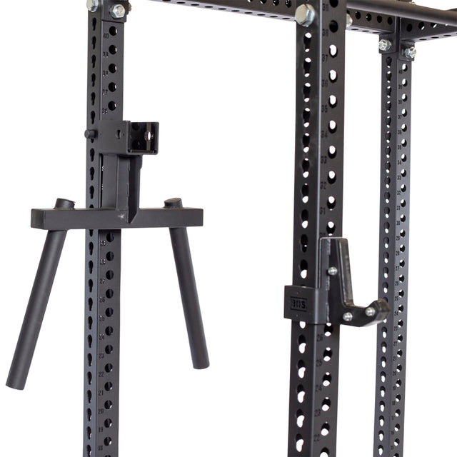 A close-up of the Bells of Steel power rack frame in black showcases multiple holes for adjustable bar placements. The rack is equipped with heavy-duty accessories including the Y Dip Bar Rack Attachment and a J-hook, both designed for strength training exercises.