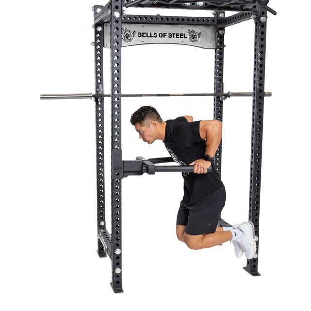 A man executes chest dips on a power rack fitted with Y Dip Bar Rack Attachments. He is dressed in a black T-shirt and shorts, complemented by white sneakers. The robust rack prominently features the "Bells of Steel" brand logo at the top.