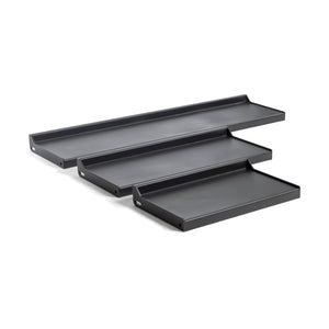 The Bells of Steel Dumbbell/Kettlebell Shelf features four black, rectangular metal shelves with raised edges and visible mounting holes, ideal for displaying collections or storing gear. These wall-mounted shelves are shown in a staggered arrangement for versatile functionality.