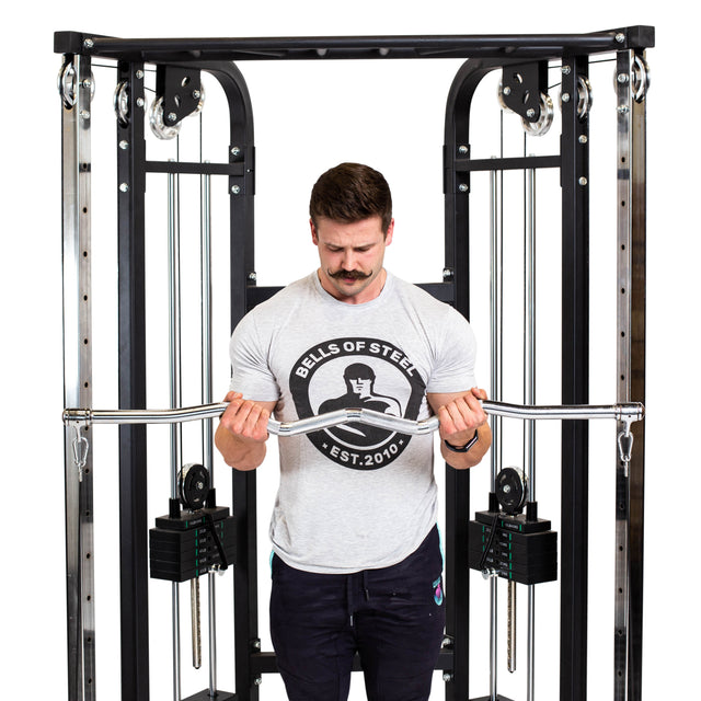 A man in a Bells of Steel t-shirt does a bicep curl with the Double Hook EZ Curl Bar Cable Attachment, positioned centrally for joint-friendly barbell exercises.