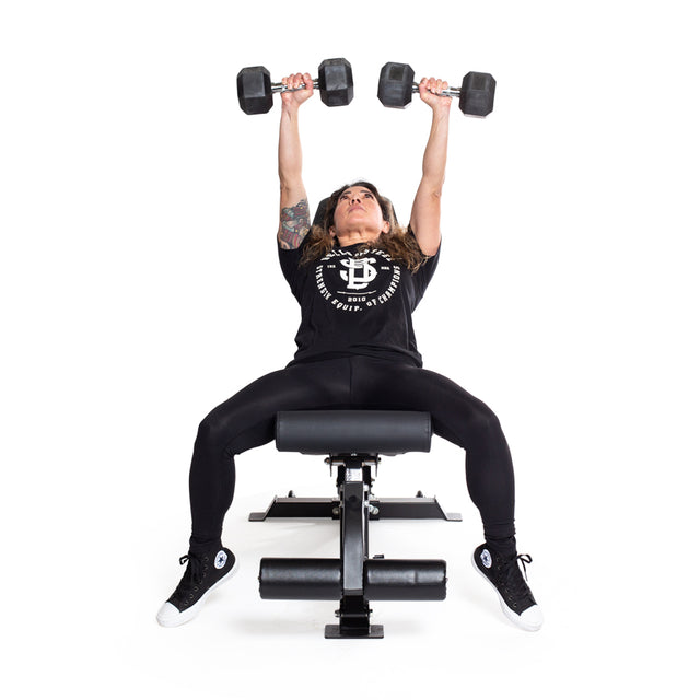 Dressed in black, a person lies on a weight bench, performing a dumbbell press with Bells of Steel's Ergo Rubber Hex Dumbbells. They extend both arms upward against a plain white backdrop.