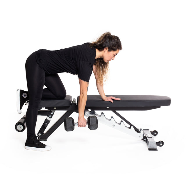 A woman in black workout attire and sneakers performs a dumbbell row on an adjustable bench, set against a white background. With one knee and hand on the bench, she uses an Ergo Rubber Hex Dumbbell by Bells of Steel featuring an ergonomic grip.