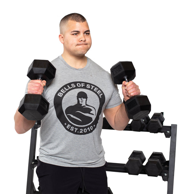 A person in a gray "Bells of Steel" t-shirt lifts two Ergo Rubber Hex Dumbbells, featuring an ergonomic grip, in front of a weight rack stocked with more dumbbells.