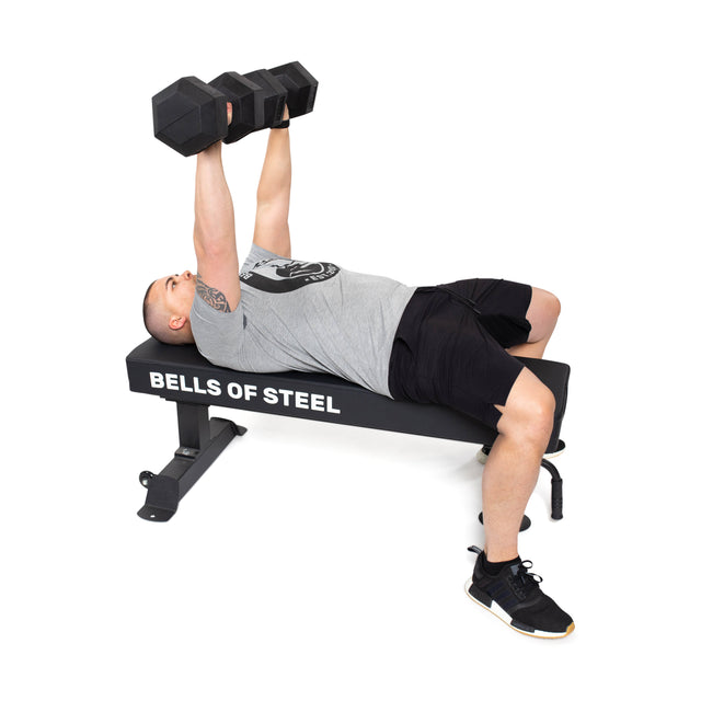 A person in a gray shirt and black shorts is lying on a "Bells of Steel" weight bench, performing a dumbbell bench press with Ergo Rubber Hex Dumbbells featuring commercial-grade chrome-plated handles.