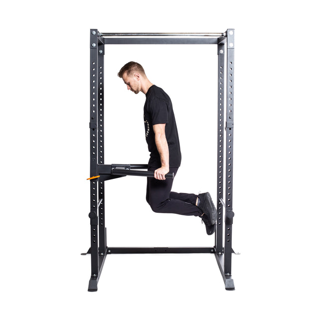 A man executes a dip exercise using the Bells of Steel Y Dip Bar Rack Attachment on a squat rack equipped with parallel dip bars. He is wearing a black shirt and pants, with the squat rack prominently displayed against the plain white background.