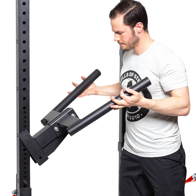 A man in a gray t-shirt is adjusting a Bells of Steel Y Dip Bar Rack Attachment on a black workout rack. Focused on securing the equipment, he ensures it's properly set up against the plain white background.