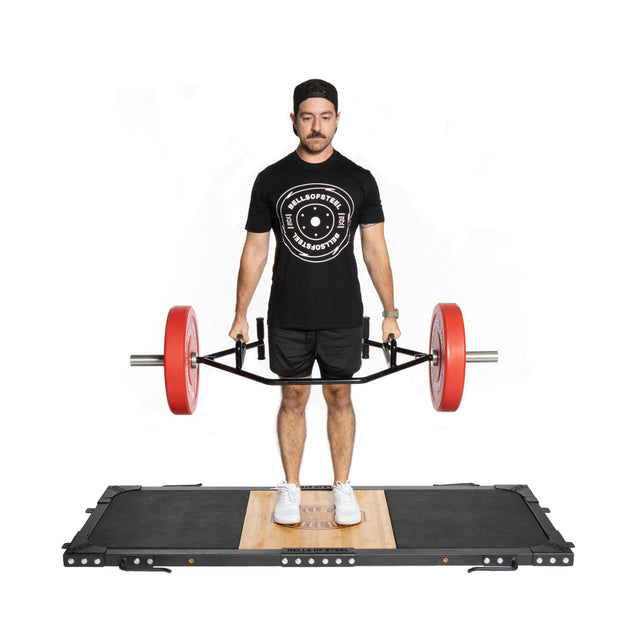 A man stands on a Bells of Steel Weightlifting Platform, hoisting an Olympic barbell with red plates. He's dressed in a black t-shirt with a circular logo, shorts, white sneakers, and a cap.