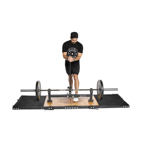 A person in athletic wear and a black cap is using the Bells of Steel Weightlifting Platform, poised with an Olympic barbell loaded with weights. The plain white background highlights their focus and determination.