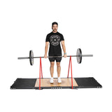 A man in a black shirt and shorts lifts an Olympic barbell on a Bells of Steel Weightlifting Platform. He wears a black cap and white sneakers. The barbell is supported by red stands, set against a plain white background.