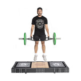 A man stands on a Bells of Steel Weightlifting Platform, lifting a trap bar with green weights. He's dressed in a black cap, logoed T-shirt, black shorts, and white sneakers against a white backdrop. The platform prominently features its logo.