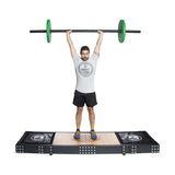 A man in a gray shirt and black shorts confidently performs an overhead lift with an Olympic barbell featuring green weights. With fully extended arms, he wears a cap and orange sneakers while standing on the Bells of Steel Weightlifting Platform.