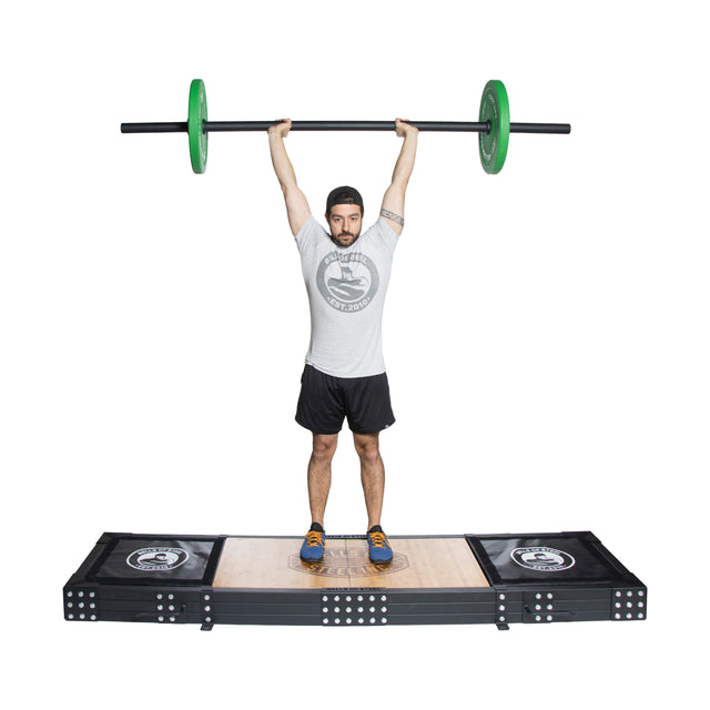 A man in a gray shirt and black shorts confidently performs an overhead lift with an Olympic barbell featuring green weights. With fully extended arms, he wears a cap and orange sneakers while standing on the Bells of Steel Weightlifting Platform.