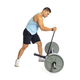 A man in a light blue tank top and black shorts uses the Deadlift Jack with Rollers by Bells of Steel for a landmine exercise, adjusting weights while leaning forward and gripping a barbell on a white background.