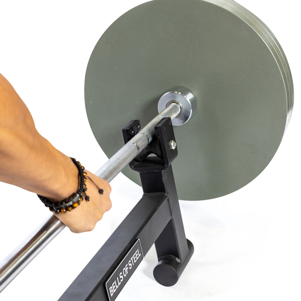 A person gripping a barbell with heavy weight plates is ready for action, supported by a Bells of Steel squat rack. Their wrist features a beaded bracelet, and seamless weight changes are aided by the Bells of Steel Deadlift Jack with Rollers against a plain white backdrop.