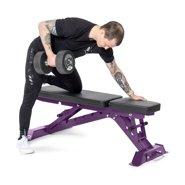 Adorned with tattoos on both arms and wearing white sneakers, a person in black performs a one-arm dumbbell row using the Buzz-Saw Heavy-Duty Adjustable Bench by Bells of Steel, positioning their right knee and left hand on the bench.