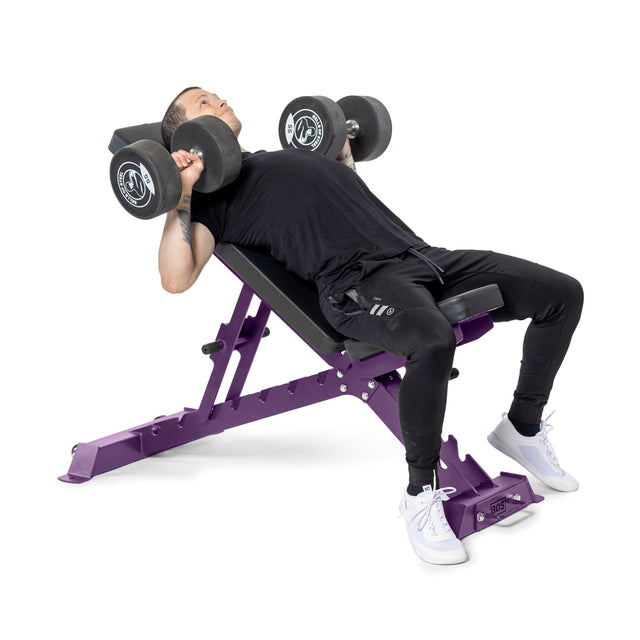 A person in a black T-shirt and pants performs an incline dumbbell bench press on the purple Buzz-Saw Heavy-Duty Adjustable Bench by Bells of Steel, holding a dumbbell in each hand, wearing white sneakers, and leaning back as part of their exercise routine.