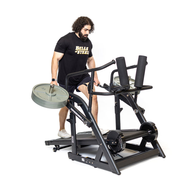 A man in a black Bells of Steel t-shirt uses "The Dreadmill," a strength-training treadmill with manual incline, magnetic resistance lever, multiple weights, and a sturdy frame.