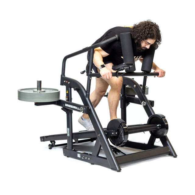 A curly-haired person uses The Dreadmill by Bells of Steel, a black manual incline treadmill with padded supports and weight plates. Positioned forward, they grip the handles against a clean white background.