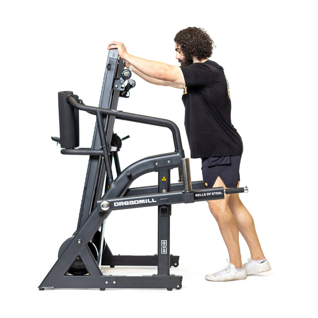 A person is using "The Dreadmill" by Bells of Steel, a manual incline treadmill. They lean forward, hands gripping the sleek black frame with a magnetic resistance lever for effective strength training.